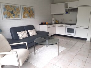 Un salon moderne avec un canapé, une table basse en verre et un coin cuisine doté de meubles blancs et d’appareils électroménagers.