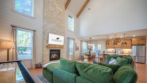 Ein geräumiges Wohnzimmer mit einem grünen Sofa, einem Kamin, einem Billardtisch und einem im Hintergrund sichtbaren Küchenbereich.
