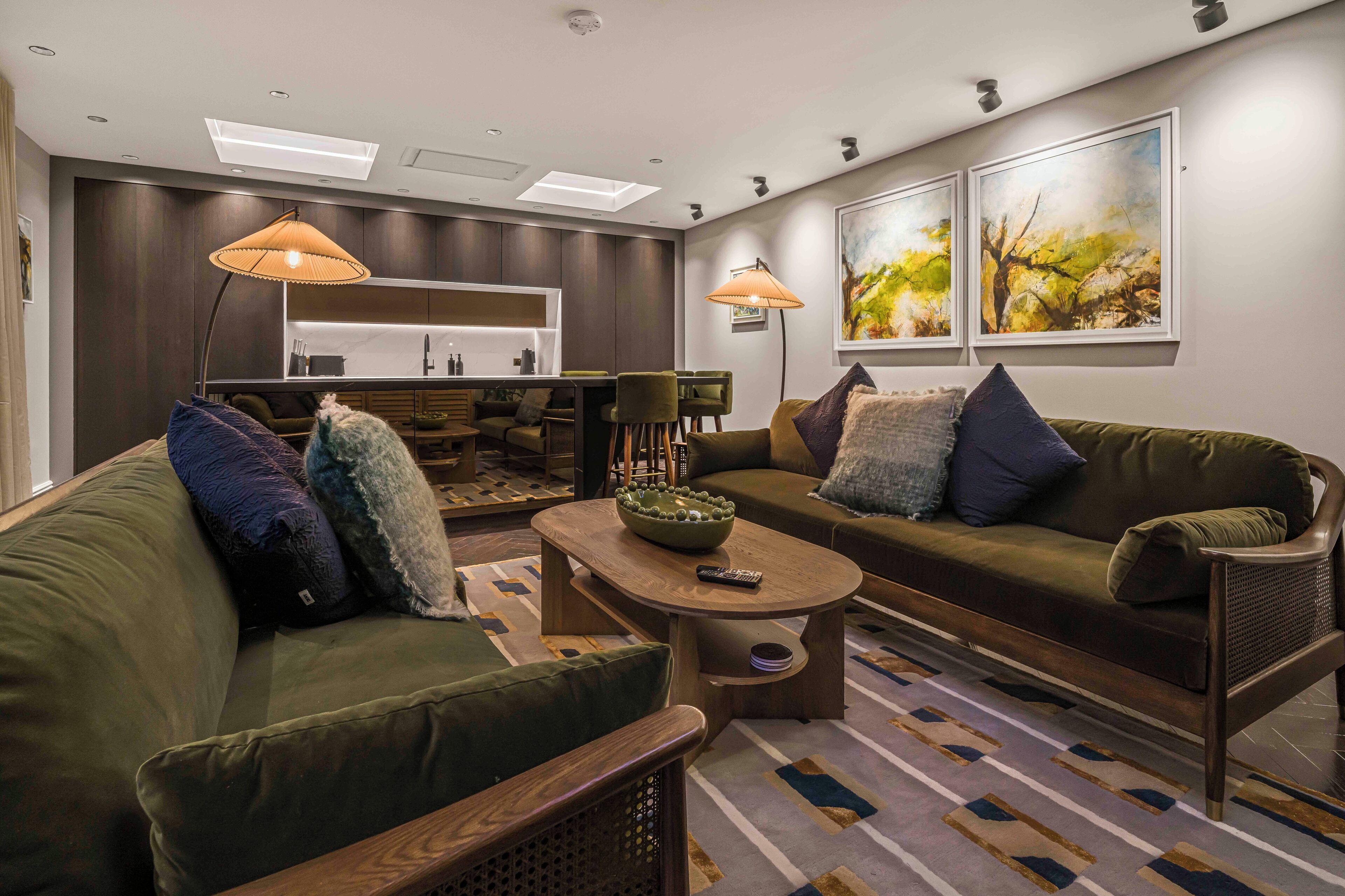 Un salon moderne avec un canapé, un fauteuil et une table basse. On aperçoit à l’arrière-plan un coin cuisine avec un bar et des sièges.