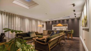 A modern living room with a sofa, dining table, and chairs. There is a kitchen area with a dining table and chairs. The room has a large window with curtains and a ceiling with recessed lighting.