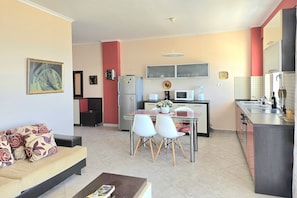 A modern kitchen with a refrigerator, microwave, and sink. There is a dining area with a glass table and white chairs. A sofa with patterned cushions is in the living area. - Dream View Apartment Durres 119 (Durrës)