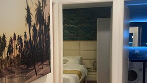 A bedroom with a bed, a nightstand, and a painting of palm trees. - Vacation Rental Apartment with Shared Garden, Balcony & Wi-Fi (Bezons)