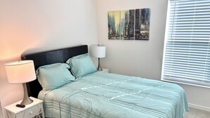 Ein ordentlich bezogenes Bett mit einer hellblauen Tagesdecke, eine Nachttischlampe und ein kleiner Nachttisch mit Fernbedienung.