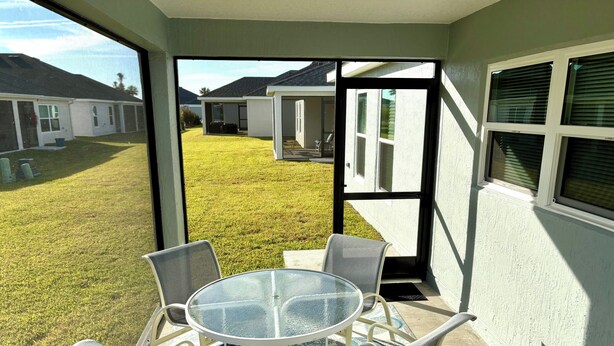 Een overdekt terras met een glazen tafel en stoelen, een gekleurd vloerkleed en uitzicht op een gazon met huizen op de achtergrond.