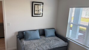 Een kleine, gezellige kamer met een grijze bank, twee kussens, een lijst met een foto aan de muur en een raam met zonwering.