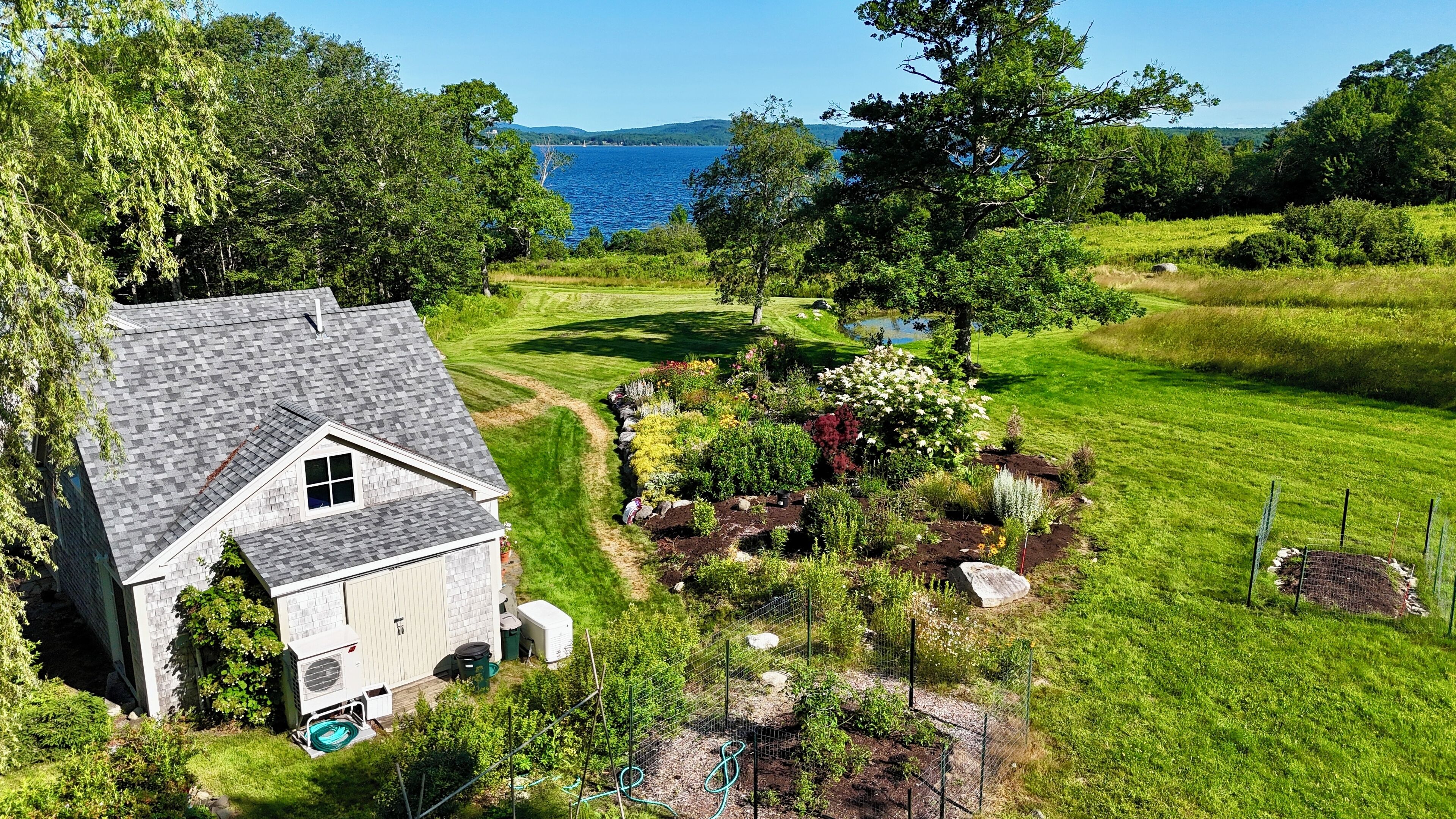 Una casa con techo inclinado, un pequeño jardín con plantas variadas y vistas a un lago.