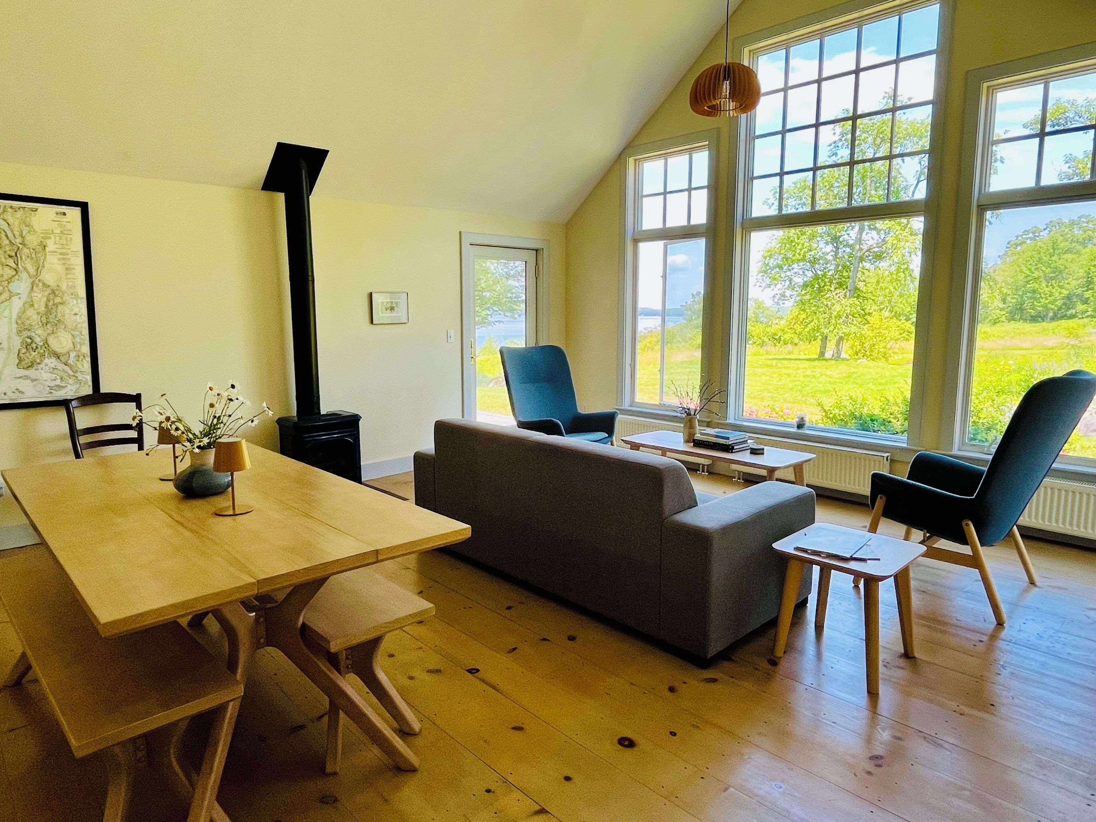 Una habitación con una mesa de madera, un sofá gris, dos sillas azules, un calentador negro y ventanas grandes con vistas a zonas verdes.