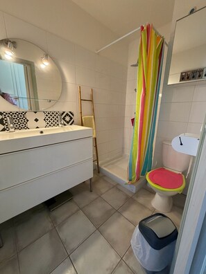 A bathroom with a white vanity, a mirror, a shower with a colorful curtain, a toilet, and a trash bin. - Studio 2OO M Centre Saint Florent -in quiet building. (Saint-Florent)