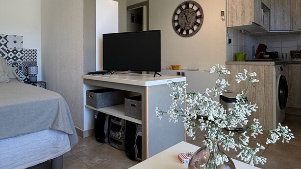 A compact living space with a bed, a flat-screen TV, a clock, and a kitchen area with wooden cabinets. - Studio 2OO M Centre Saint Florent -in quiet building. (Saint-Florent)