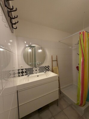 Un baño con tocador blanco, un espejo, ducha con cortina colorida y piso de baldosas.