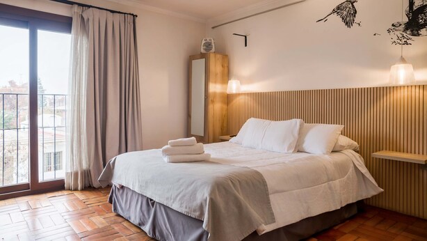 A bedroom with a bed, a bedside table, a nightstand, a window with curtains, and a wall-mounted light fixture. - Trendy Design Studio Vaisse | Modern Santiago Stay (Providencia)