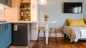 A compact kitchen with a refrigerator, a coffee maker, and a plant on the counter. There is a bed with yellow pillows and a TV mounted on the wall. - Trendy Design Studio Vaisse | Modern Santiago Stay (Providencia)