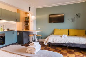 Habitación de hotel pequeña con kitchenette, una cama con almohadas amarillas, un televisor montado en la pared y un mostrador de recepción.