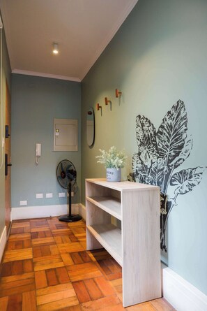 Un pasillo con suelo de madera, un ventilador de pared, una planta en maceta y un estante con un espejo.