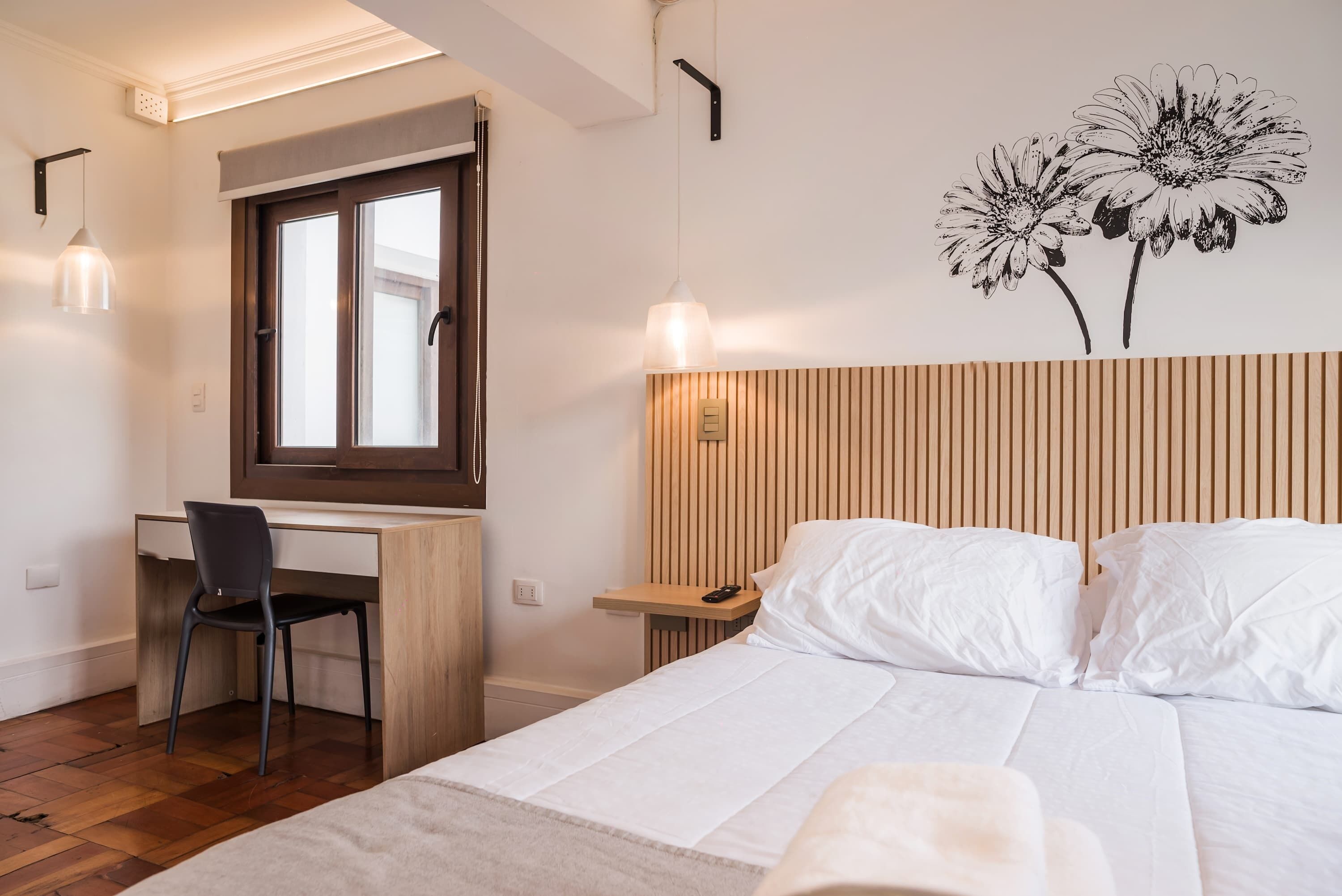 A hotel room with a bed, a desk, a chair, a window, and a wall-mounted light fixture.
