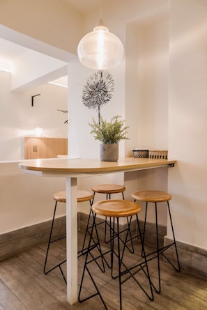 A modern kitchen with a wooden table, four stools, a hanging light fixture, and a potted plant.