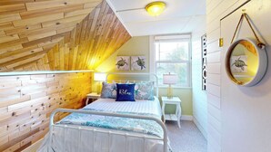 Ein Schlafzimmer mit schrägem Dach, Holzvertäfelung, einem Bett mit weißem Metallgestell, einem Nachttisch mit Lampe und einem Fenster mit Blick ins Grüne.