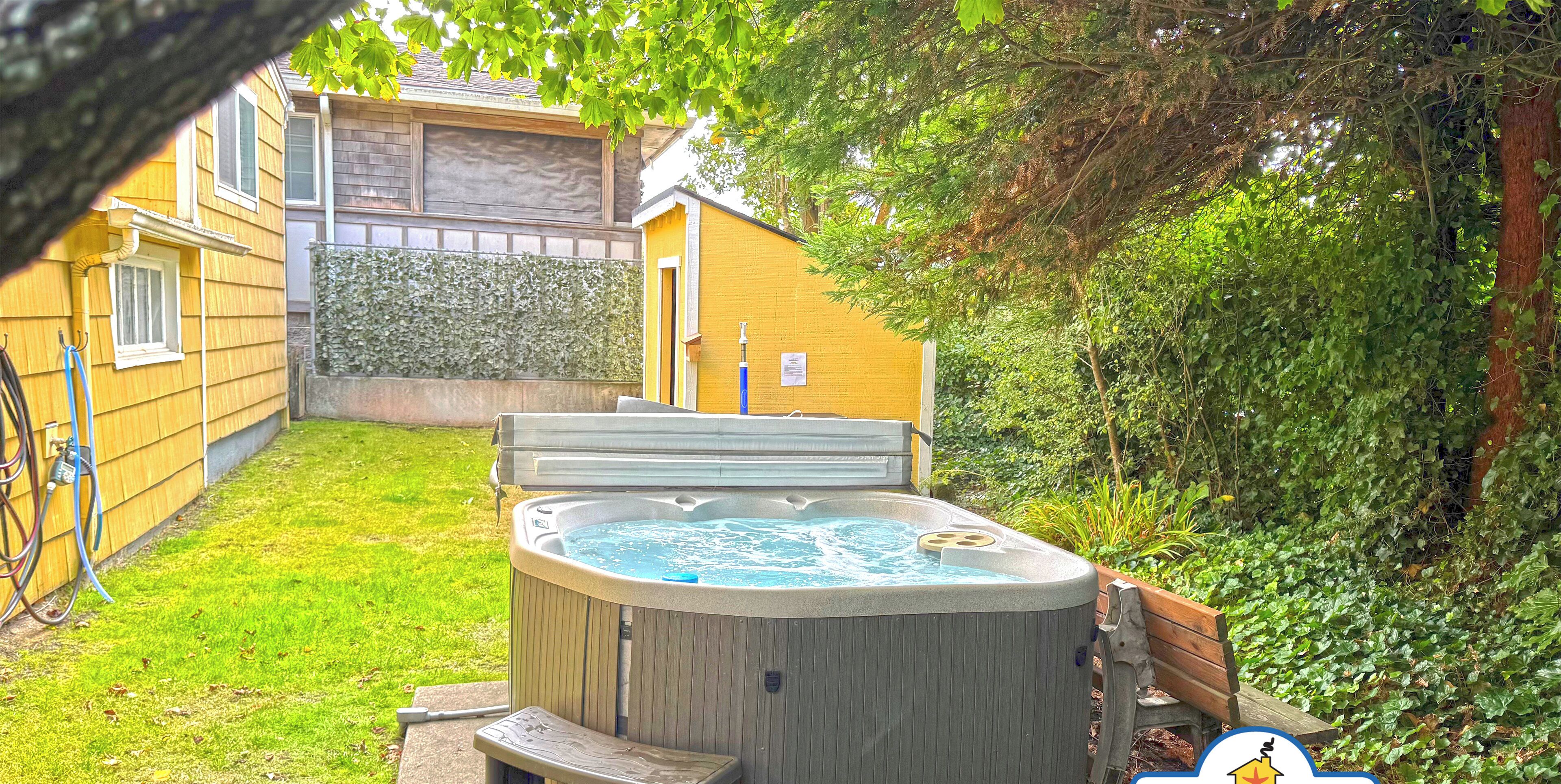 A hot tub in a backyard with a wooden deck, a hedge, and a house in the background.