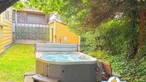 Ein Whirlpool in einem Garten mit Holzterrasse, Hecke und einem Haus im Hintergrund.