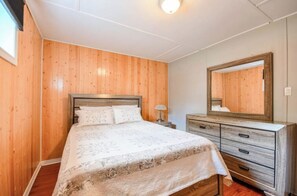 A bedroom with a wooden wall, a bed with a patterned bedspread, a dresser with drawers, and a mirror above the dresser.