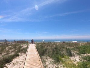 En träpromenad som leder till en strand med gles vegetation och en klar, blå himmel.