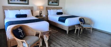 A room with two beds, a chair, a small table, and a lamp. There are two framed pictures on the wall.