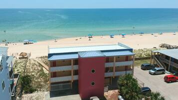A beachfront hotel with a red building, a parking area, and a sandy beach with umbrellas.