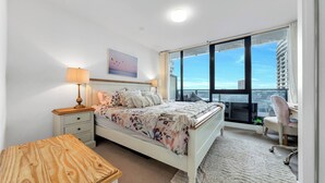 Ein modernes Schlafzimmer mit einem großen Bett, einem Nachttisch mit Lampe, einem Schreibtisch mit Stuhl und Blick auf die Stadt durch große Fenster.