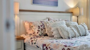 Ein ordentlich bezogenes Bett mit floralem Bettbezug und dekorativen Kissen, ein hölzernes Kopfteil und ein an der Wand hängendes Bild von Vögeln.