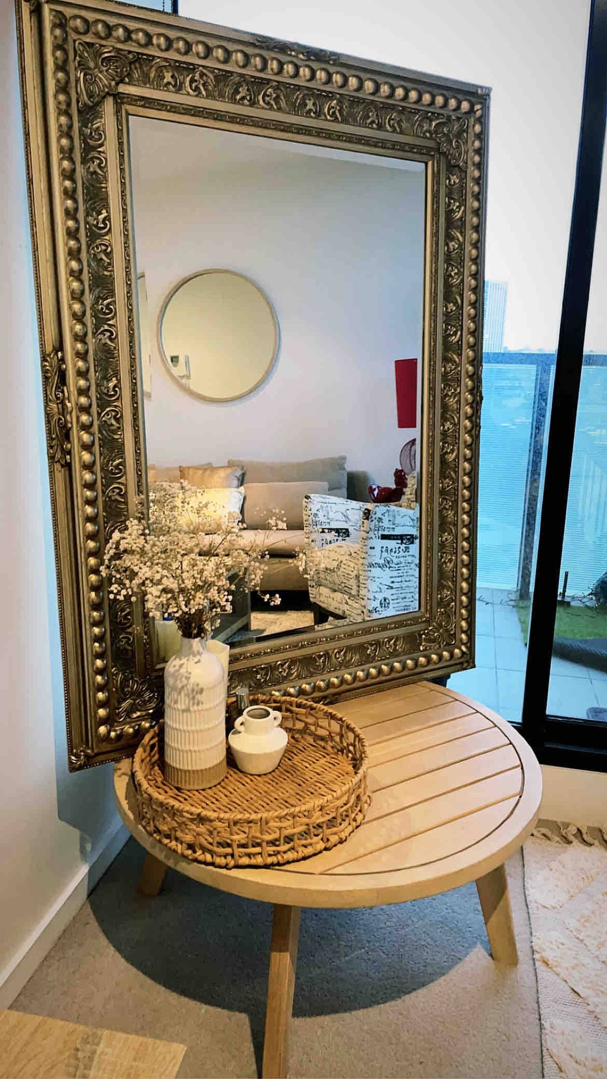 A large ornate mirror with a decorative frame, a round table with a woven basket and a vase of flowers, and a view of a bedroom with a bed and dresser.