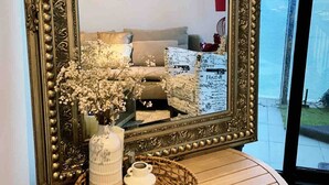 Ein großer, aufwendig gestalteter Spiegel mit dekorativem Rahmen, ein runder Tisch mit einem gewebten Korb und einer Blumenvase, sowie ein Blick auf ein Schlafzimmer mit Bett und Kommode.