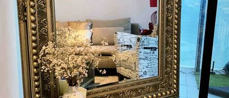 A large ornate mirror with a decorative frame, a round table with a woven basket and a vase of flowers, and a view of a bedroom with a bed and dresser.