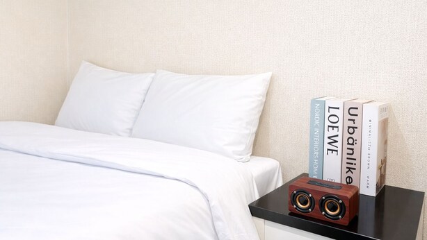 Una cama bien hecha con ropa de cama blanca, una mesita de noche negra con un altavoz y tres libros titulados 'Nordic Interiors Home', 'LOEWE' y 'Urban Like'.