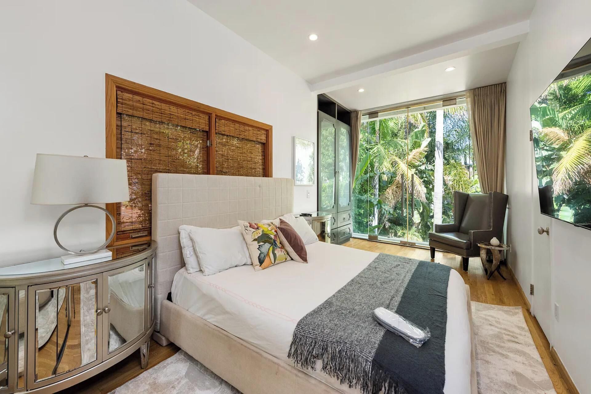 A modern bedroom with a large bed, a bedside table, a lamp, a chair, and a view of greenery through large windows.