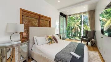 A modern bedroom with a large bed, a bedside table, a lamp, a chair, and a view of greenery through large windows.