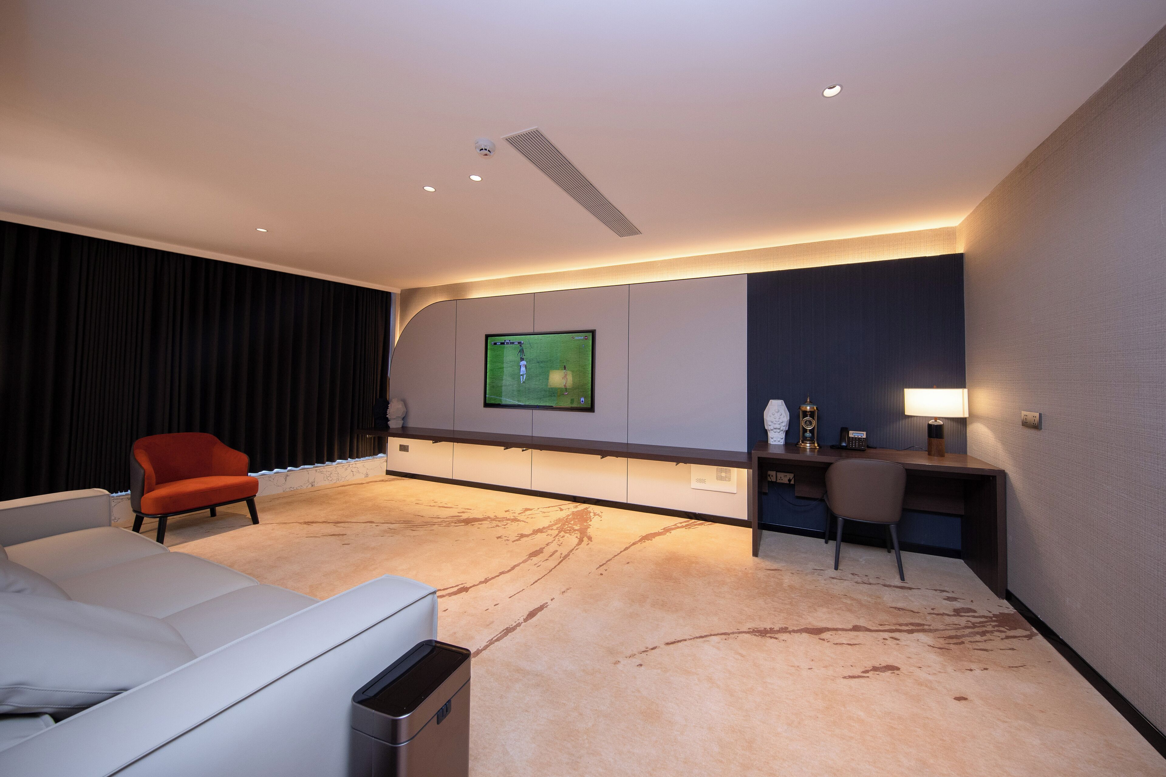 A modern hotel room with a large flat-screen TV mounted on the wall, a white sofa, a red armchair, a desk with a lamp, and a trash bin.