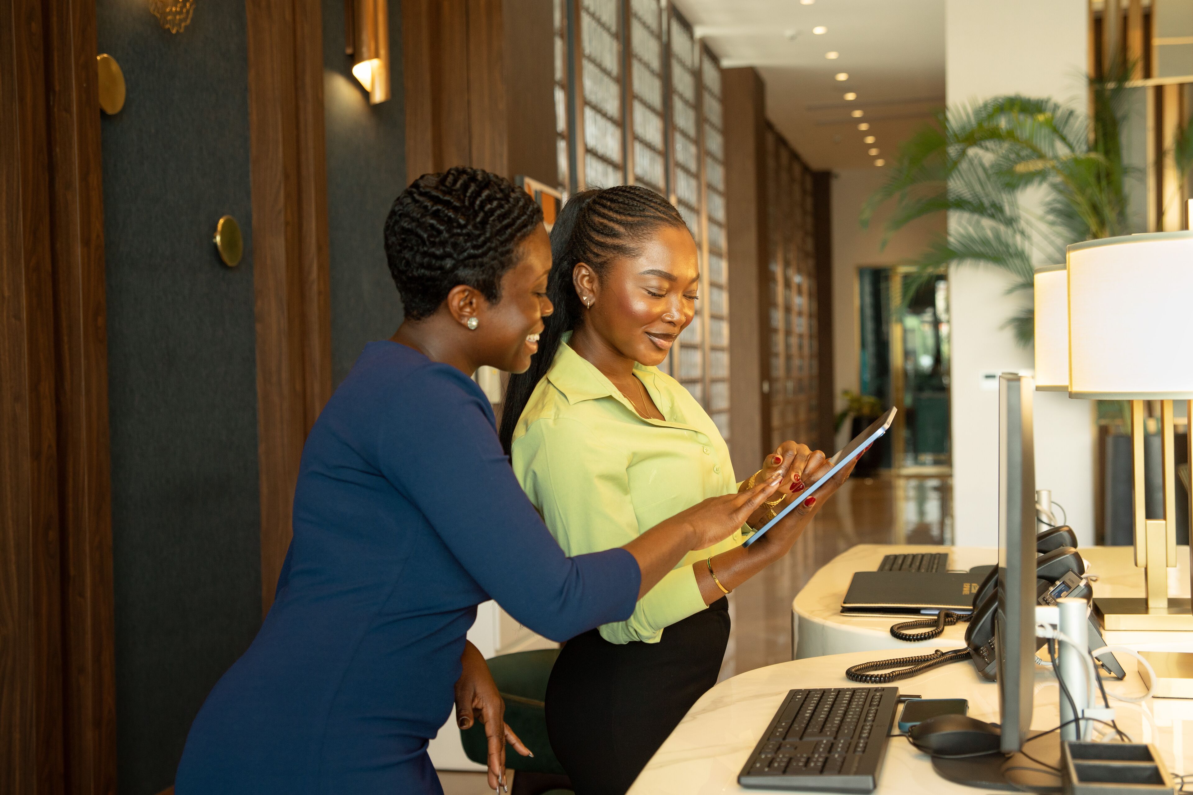 Two individuals at a reception desk with a computer, phone, and desk lamp.