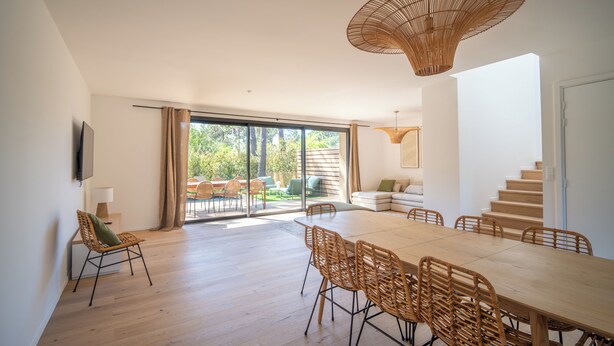 A spacious dining area with a large table and chairs, a wicker chair with a cushion, a hanging light fixture, and a view of the outdoors through large windows. - Villa Benedettu (Lecci)