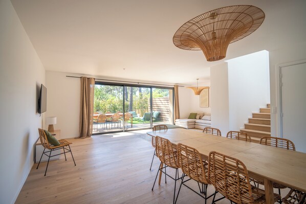 A spacious dining area with a large table and chairs, a wicker chair with a cushion, a hanging light fixture, and a view of the outdoors through large windows. - Villa Benedettu (Lecci)