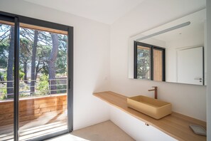 A modern bathroom with a wooden vanity, a large mirror, and a view of trees through a sliding door. - Villa Benedettu (Lecci)