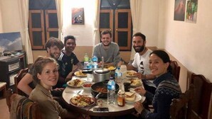 Un groupe de personnes est assis autour d’une table ronde, devant divers plats, dans une pièce dotée de fenêtres en arc de cercle et où un téléviseur est posé sur un meuble.