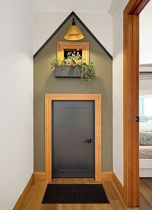 Una puerta gris oscuro con marco de madera, un picaporte negro y un felpudo negro. Sobre la puerta, una pared triangular con moldura de madera y una lámpara colgante.