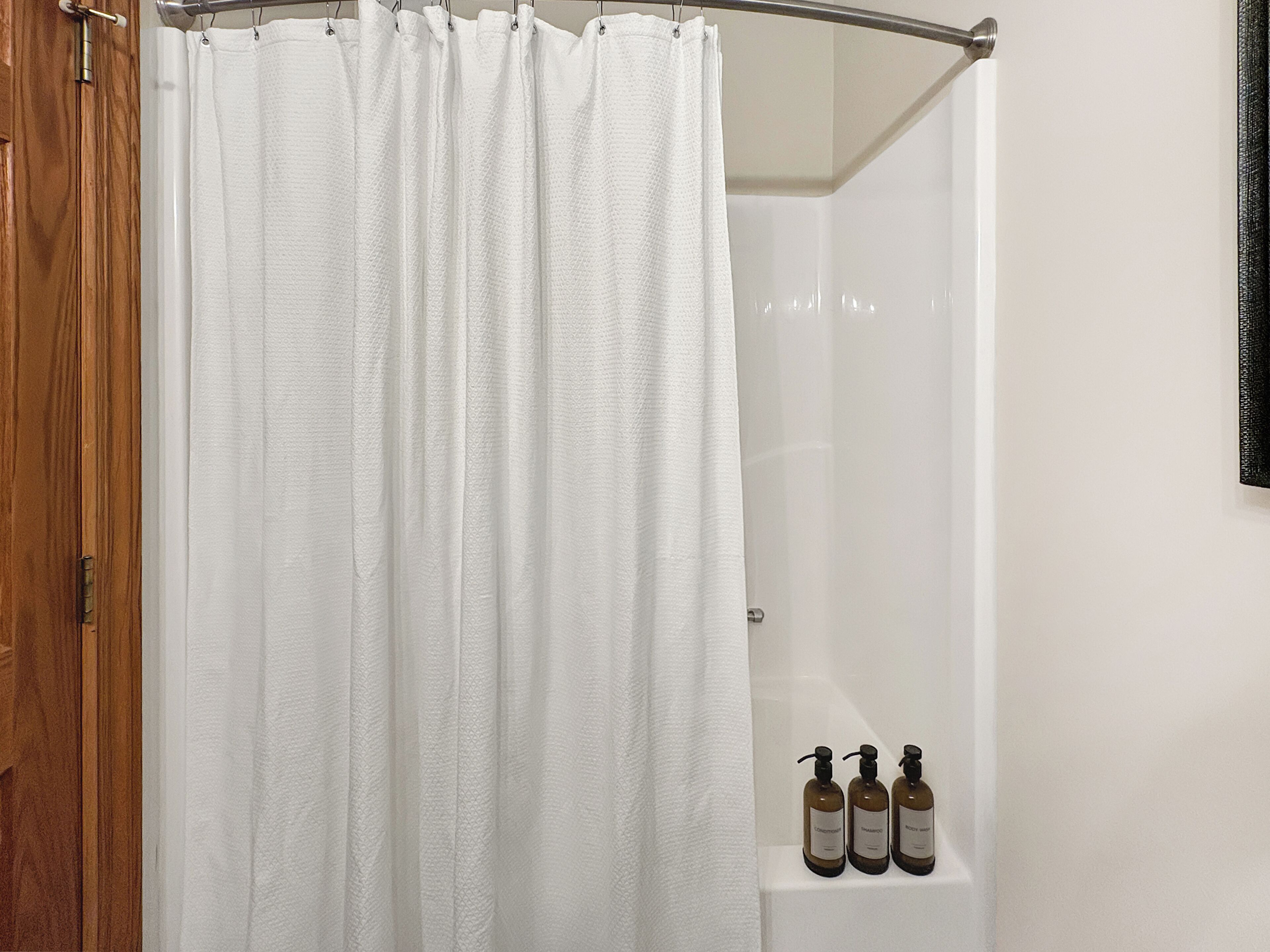 A shower area with a white shower curtain and three bottles of shampoo on a shelf.