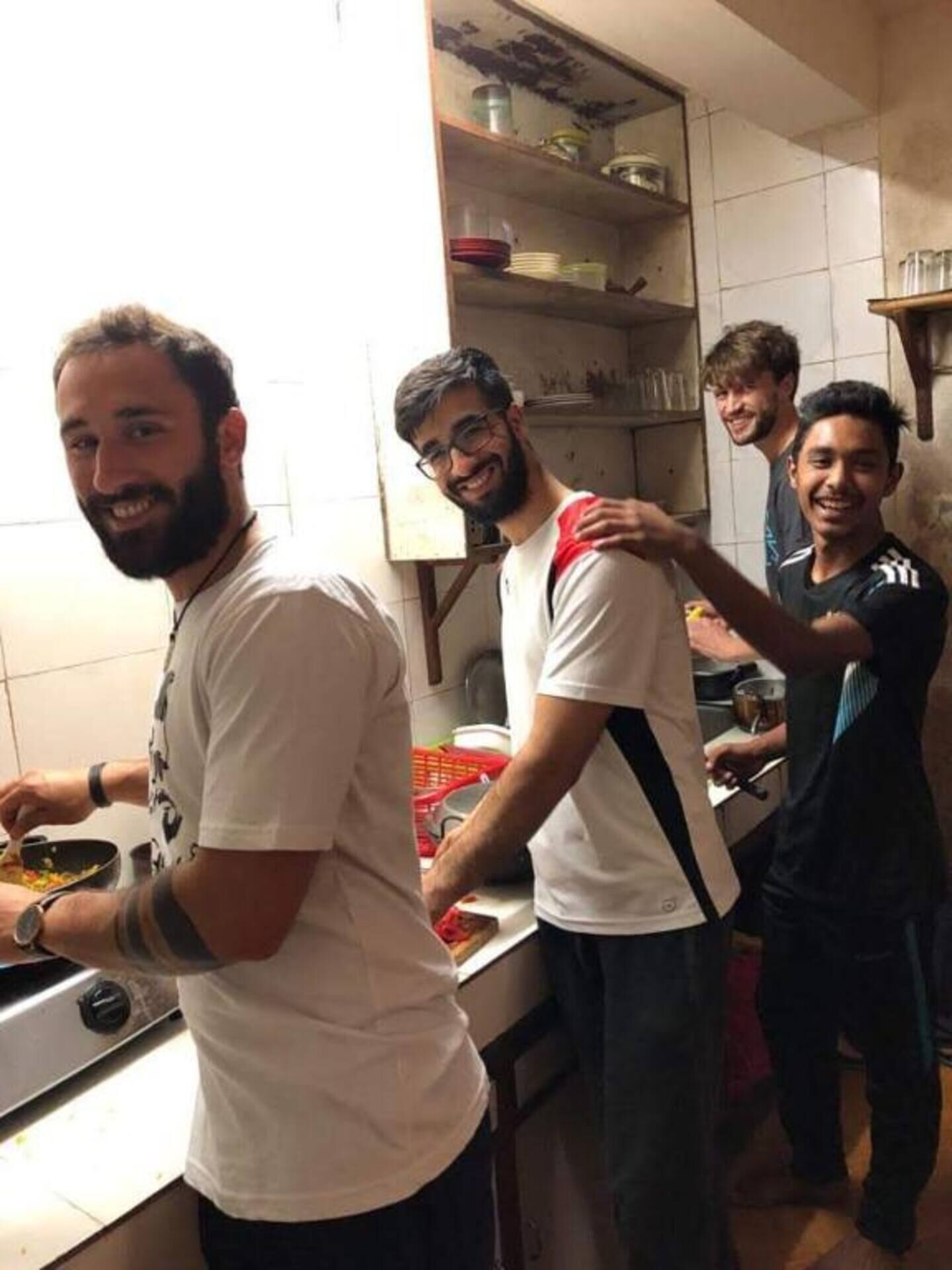 Un groupe de personnes se trouve dans une cuisine aux murs recouverts de carreaux blancs et dotée d’étagères au-dessus du plan de travail.