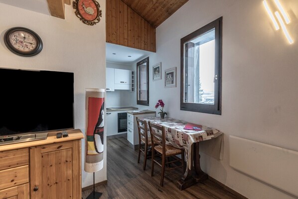 Una cocina compacta con armarios de madera, una mesa de comedor con sillas, un televisor y un reloj de pared.
