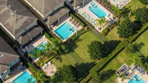 Une vue aérienne d’un quartier résidentiel avec plusieurs piscines, chacune entourée de chaises longues et de parasols, ce qui suggère un complexe hôtelier ou un lieu de vacances.