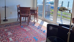 Een kamer met een groot raam, een plafondventilator, een eettafel met stoelen, een kamerbreed tapijt en een vloerlamp.