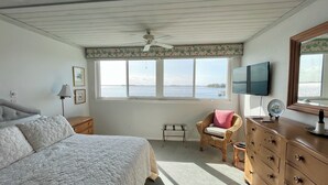 Een slaapkamer met een bed, een stoel, een dressoir, een televisie en een raam met uitzicht op zee.
