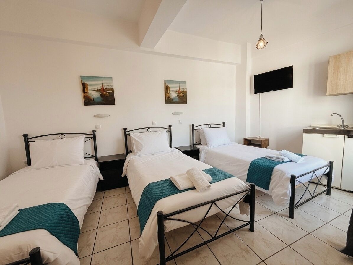 A hotel room with three single beds, a small desk with a chair, a flat-screen TV mounted on the wall, and a sink in the corner.