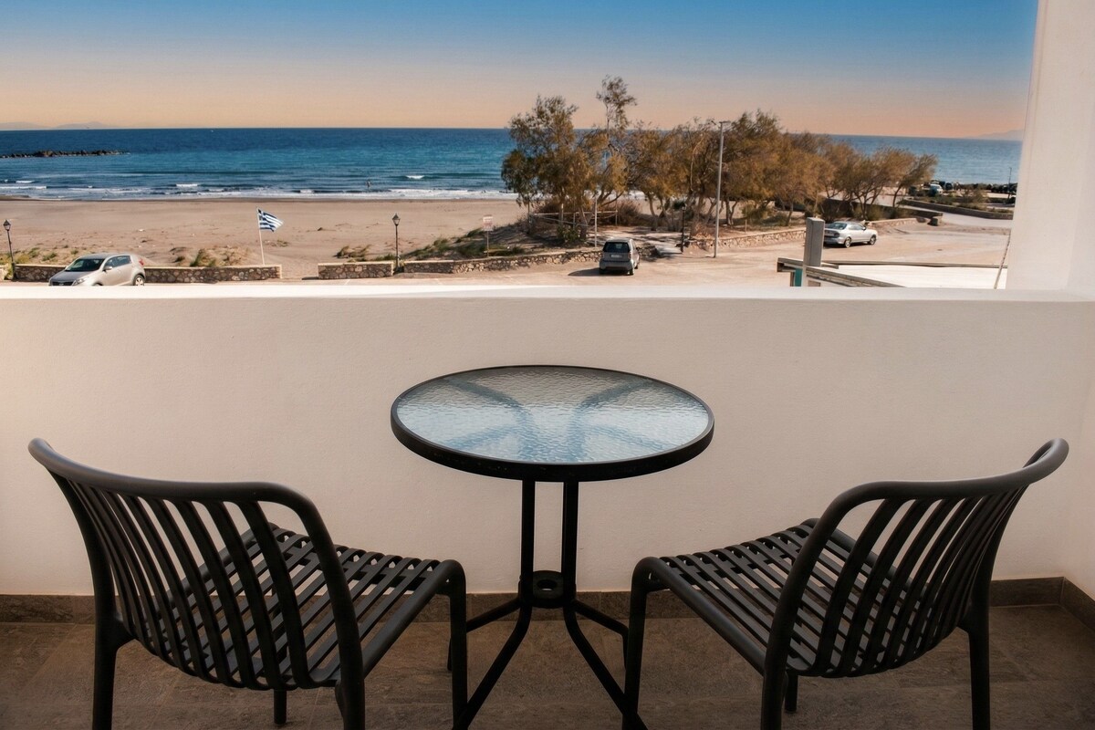 Un balcon avec deux chaises et une table, donnant sur une plage où l’on aperçoit des voitures et un drapeau.